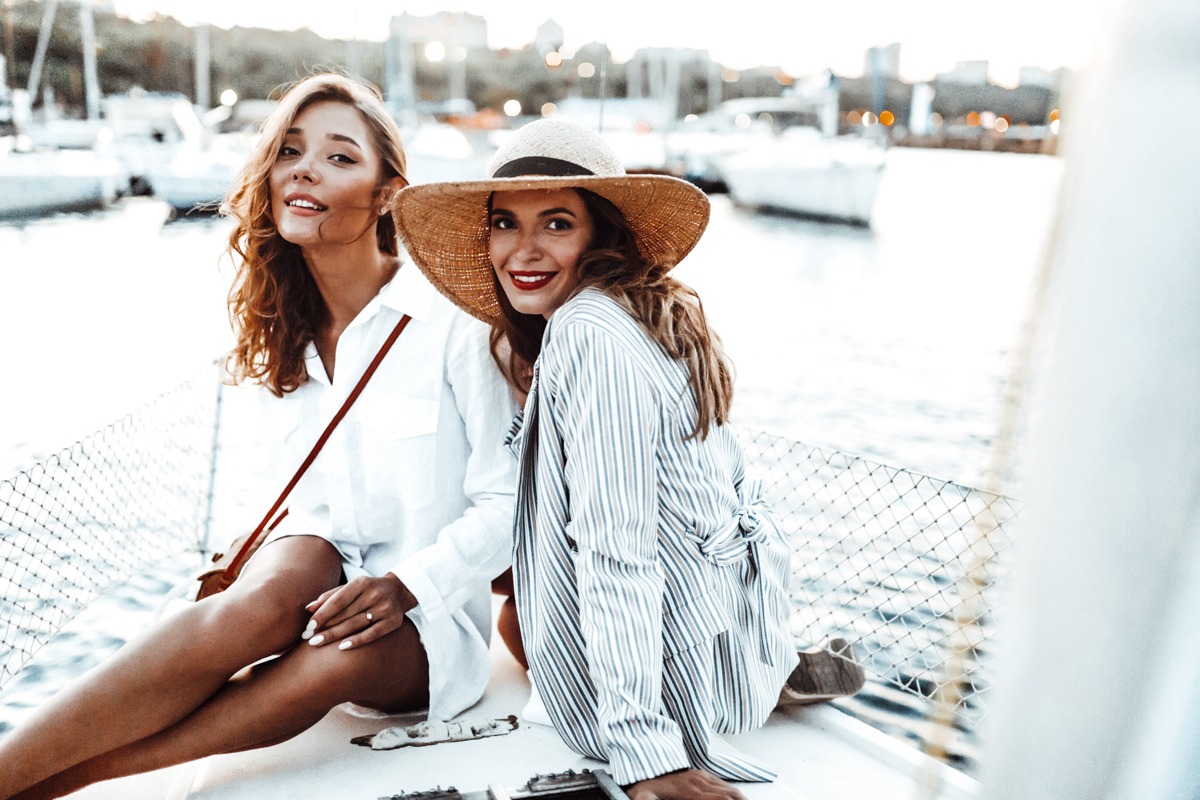 Young attractive woman in white shirt and beautiful lady in stripped jacket and straw hat dreamily looking in camera spending time together on yacht with beautiful harbor view on background
